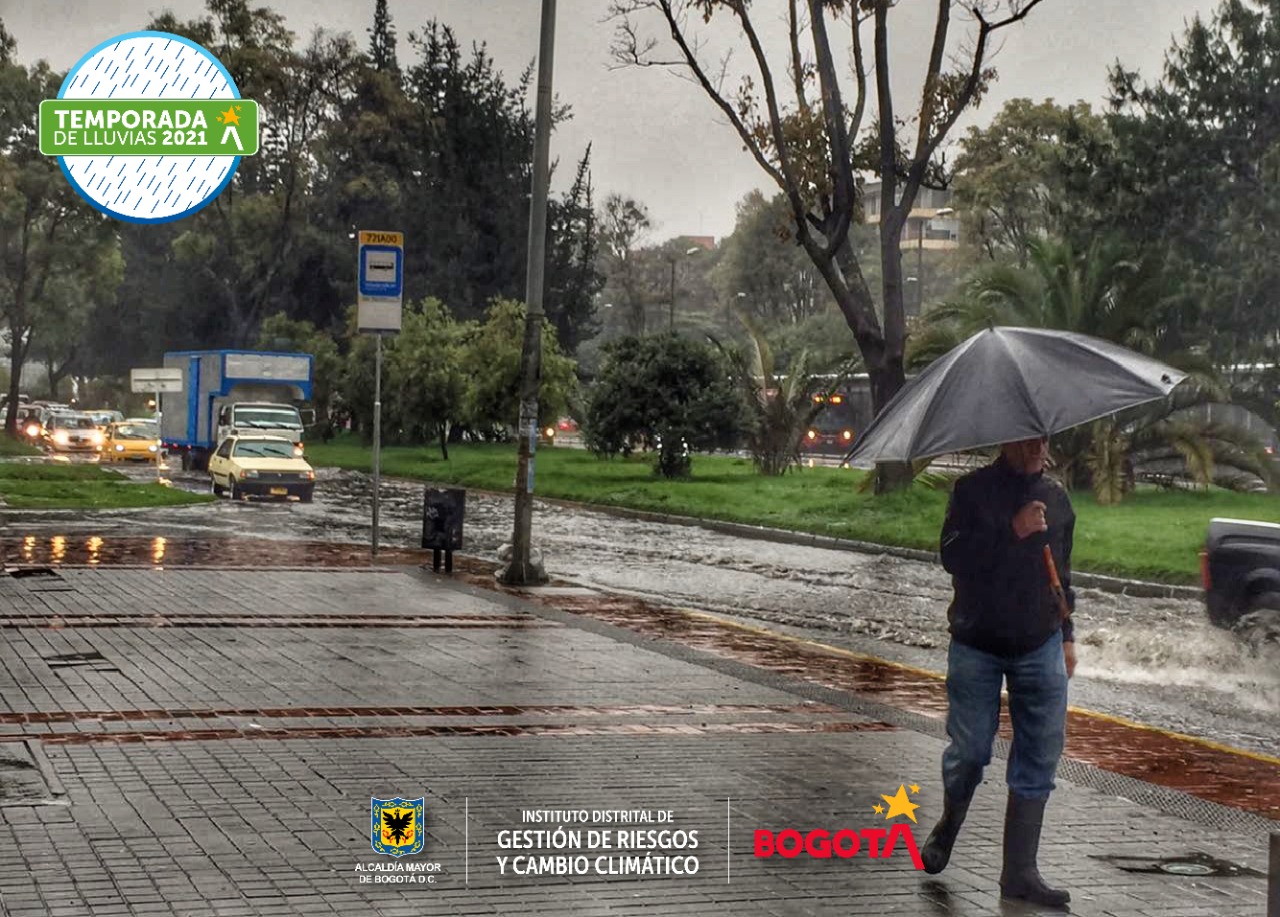 Persona con sombrilla y carros pasando por una vía co encharcamientos